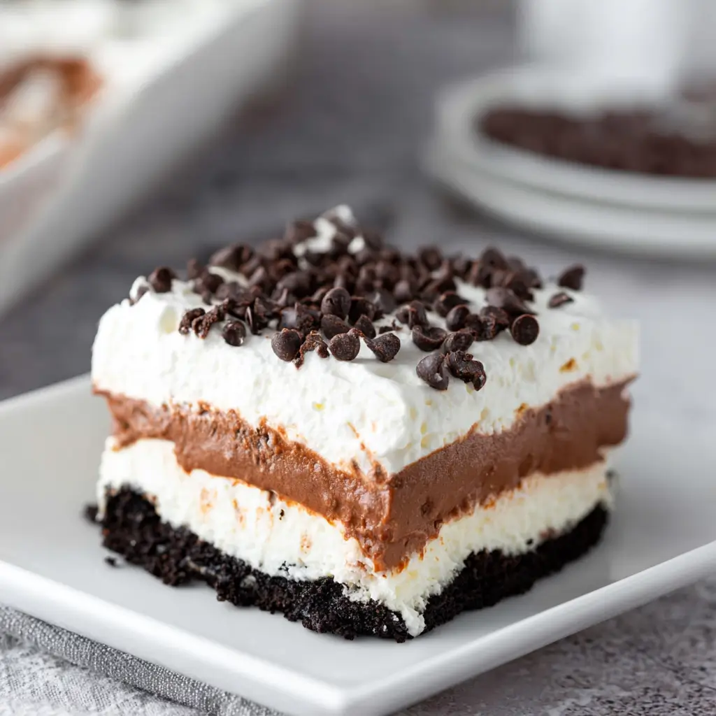 Layered chocolate lasagna dessert with Oreo crust, chocolate pudding, cream layers, and chocolate chip topping on a plate