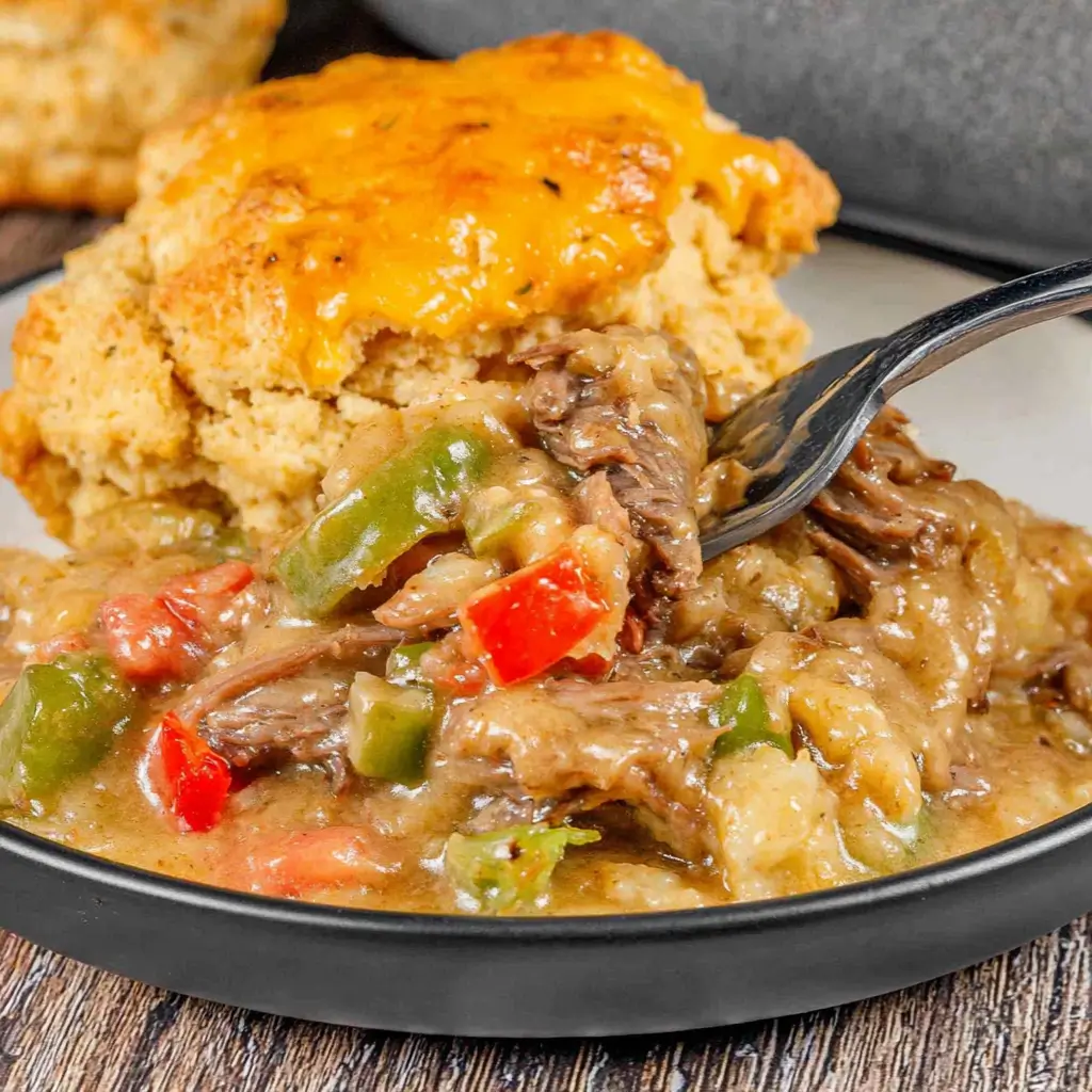A close-up of a savory beef stew with vegetables like green and red bell peppers in a thick, hearty gravy, served with a piece of fluffy cornbread topped with melted cheddar cheese.