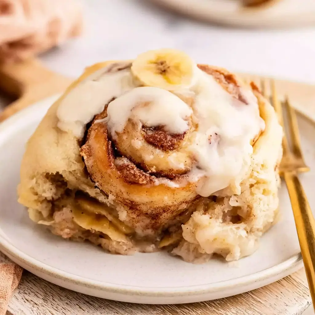 Single banana cinnamon roll on a white plate drizzled with thick cream cheese icing and topped with a fresh banana slice showing its soft swirled cinnamon layers.