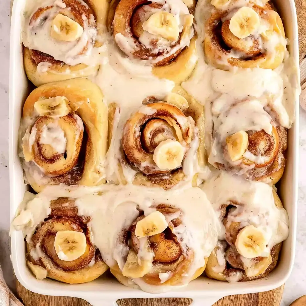 Banana cinnamon rolls baked in a white dish swirled with cinnamon sugar generously drizzled with creamy white icing and topped with fresh banana slices.