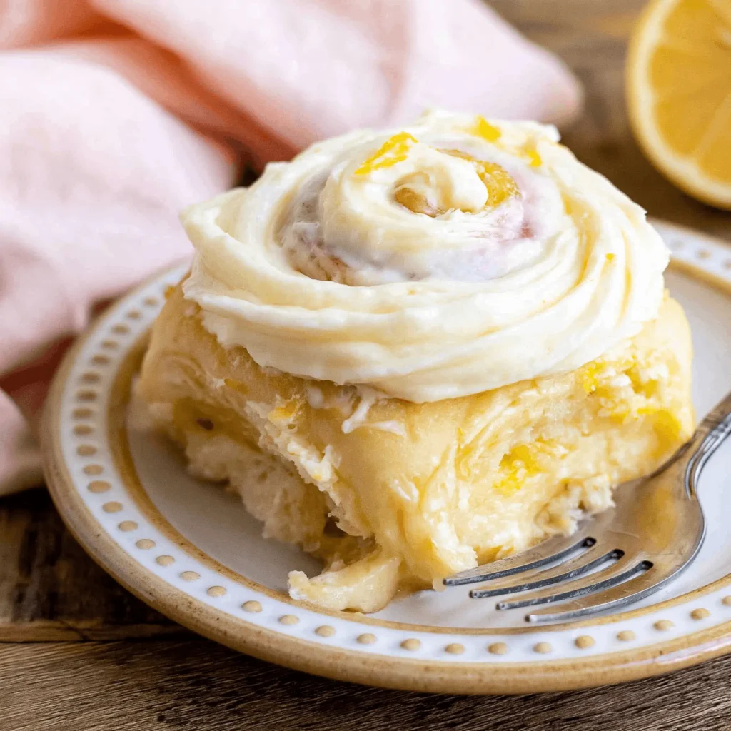 A single plated lemon roll with a fork pressed into its soft pulled apart layers, piled high with thick cream cheese frosting and scattered lemon zest on a dotted ceramic plate.