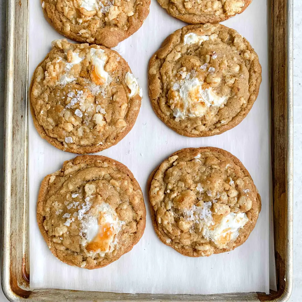 Six golden brown cookies studded with crispy rice cereal and gooey toasted marshmallow centers, finished with flaky sea salt on a parchment lined baking sheet.